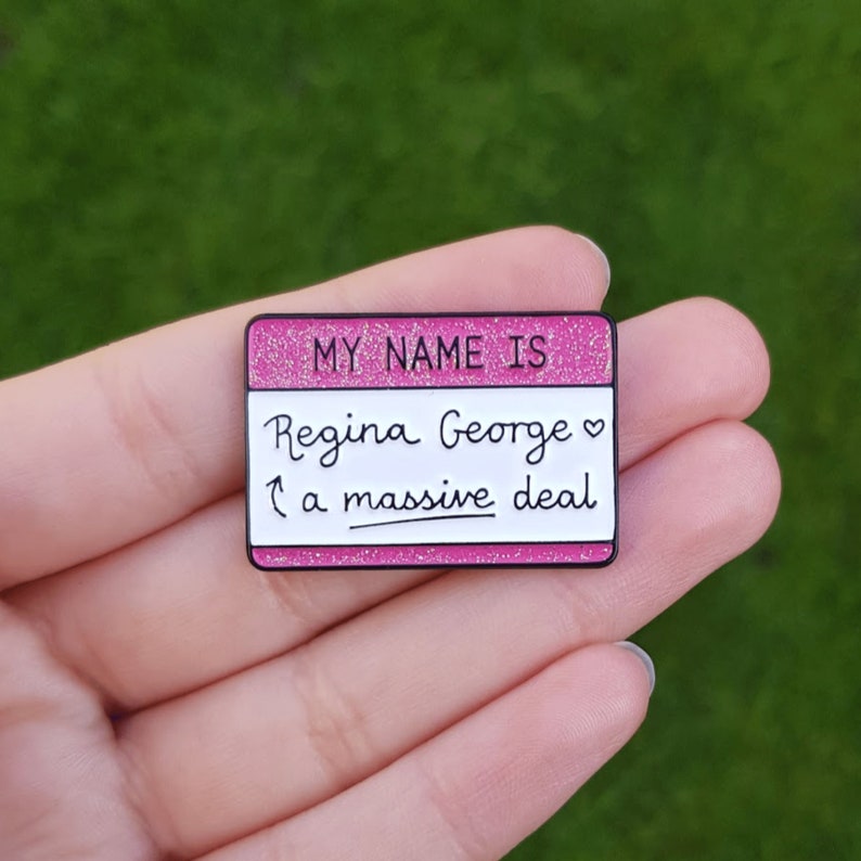 K&ouml;nnte beinhalten: Rosa glitzernder Emaille-Pin mit dem Text "My name is Regina George  a massive deal" in Wei&szlig;.