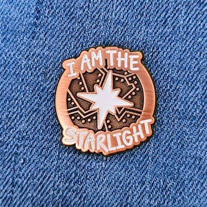 Op de afbeelding: Een koperkleurige emaille pin met de tekst "I AM THE STARLIGHT" in witte letters. De pin heeft een witte ster in het midden, omringd door een gedetailleerd ontwerp. De pin is geplaatst op een blauwe denim achtergrond.