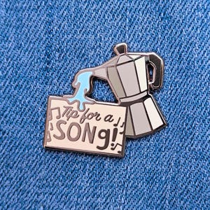May include: A silver enamel pin depicting a coffee pot pouring blue liquid onto a sign with the text "Tip for a SONG!" in a stylized font. Musical notes are on the sign. The pin is set against a blue denim background.