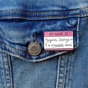 K&ouml;nnte beinhalten: Rosa emaillierte Anstecknadel mit wei&szlig;em Rand und Glitzer. Die Anstecknadel sagt "My name is Regina George &hearts; a massive deal".