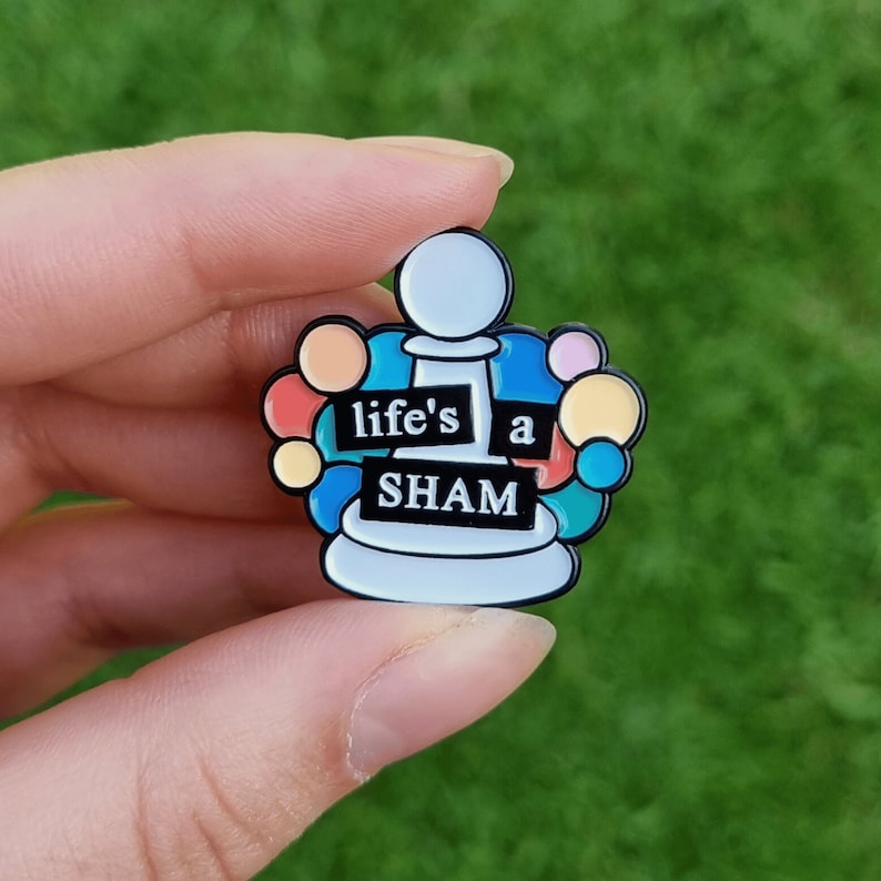 May include: A white enamel pin with a chess piece design and colorful circles around it. The chess piece has the text "life's a SHAM" on it.