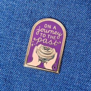 May include: A purple enamel pin with gold edging, featuring the text "ON A journey TO THE past". The pin depicts a detailed object held by two hands. The pin is set against a blue denim background.