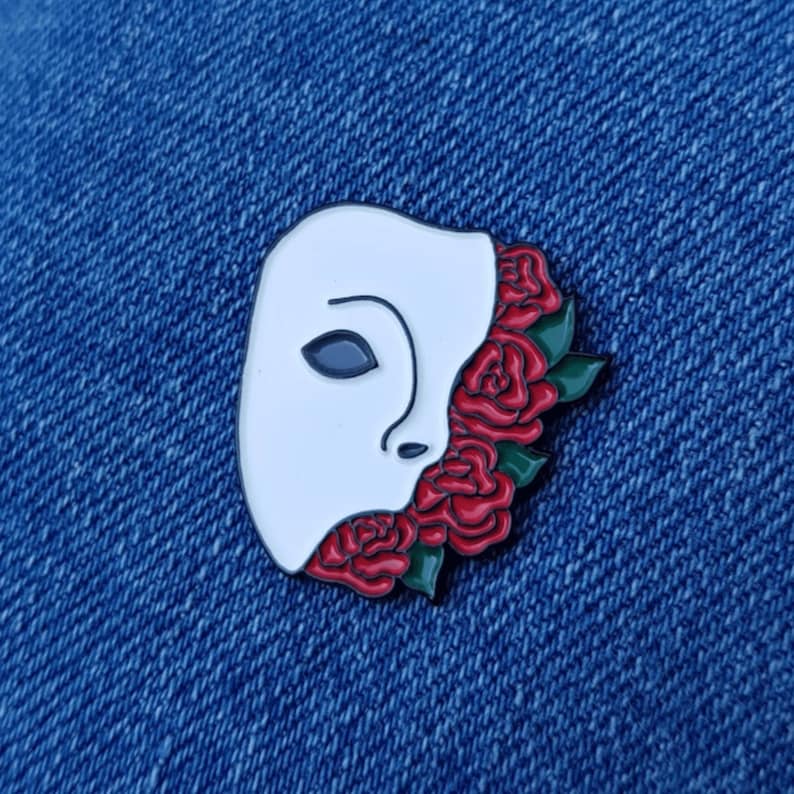 May include: White enamel pin of a face wearing a mask with red roses covering the right side of the face.