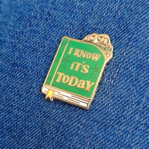 May include: A green enamel pin in the shape of a book with the words "I KNOW IT'S TODAY" in gold lettering. The pin has a gold border and a yellow bookmark. The pin is set against a blue denim background.