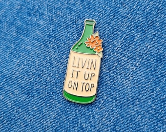 Puede incluir: Pin de esmalte verde en forma de botella con una flor en la parte superior. La botella tiene una etiqueta blanca con el texto "Livin It Up On Top".
