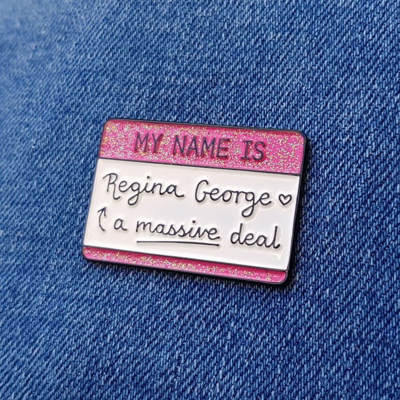 K&ouml;nnte beinhalten: Rosa-wei&szlig;er Emaille-Pin mit Glitzer. Der Pin ist in Form eines Namensschildes und sagt "My name is Regina George  &hearts;  a massive deal".