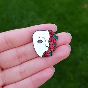 May include: White porcelain mask with a red rose design. The mask is half-covered with red roses and green leaves. The mask is a popular symbol of the Phantom of the Opera.