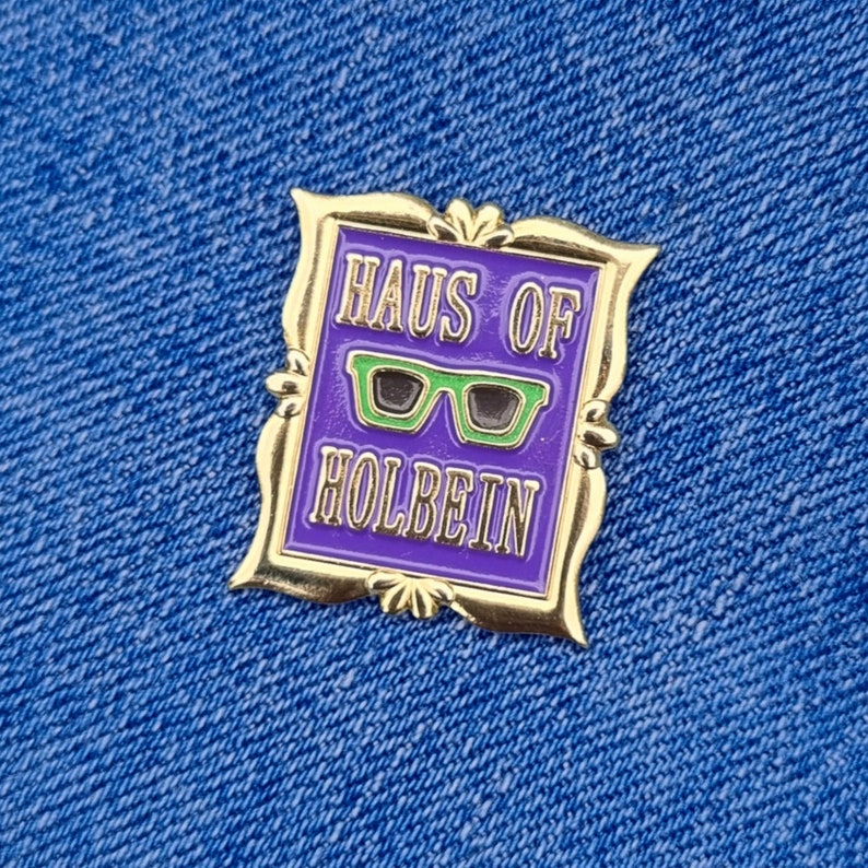 May include: A gold-colored pin with a purple background and the words "HAUS OF HOLBEIN" in gold lettering. The pin features a pair of green glasses in the center. The pin is set against a blue denim background.