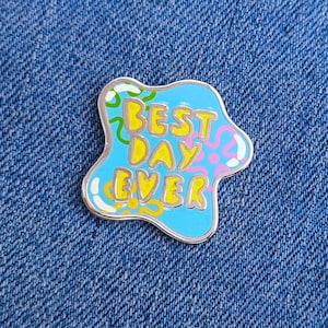 May include: A blue enamel pin with the text "BEST DAY EVER" in yellow, outlined in silver. The pin has a unique, irregular shape with colourful accents of green, pink, and yellow. The pin is set against a blue denim background.