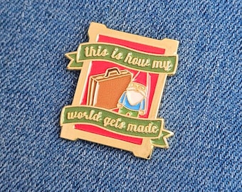 May include: A gold-colored enamel pin with a red and green design. The pin features a brown suitcase and a gnome with the text "this is how my world gets made."