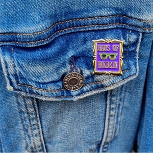 May include: A gold-framed enamel pin with the text "HAUS OF HOLBEIN" in white and purple. The pin features green sunglasses on a purple background. The pin is attached to a blue denim jacket.
