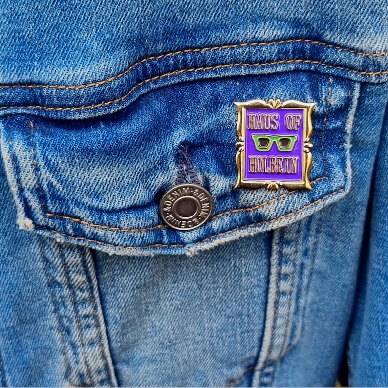 May include: A gold-framed enamel pin with the text "HAUS OF HOLBEIN" in white and purple. The pin features green sunglasses on a purple background. The pin is attached to a blue denim jacket.