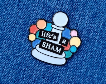 May include: A white enamel pin with a colourful design. The pin features a chess piece with the words "life's a sham" on it.
