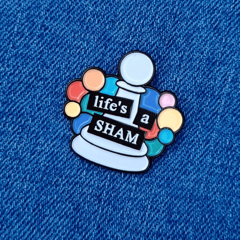 May include: A white enamel pin with a colorful design. The pin features a chess piece with the words "life's a sham" on it.