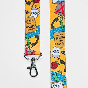 Op de afbeelding: Een gele lanyard met een herhalend patroon van een koffiemok, een telefoon, een hagedis, een roos en een bord met de tekst "We are the masters". De lanyard heeft een zilveren clip.