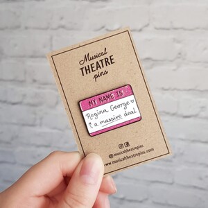 K&ouml;nnte beinhalten: Rosa emaillierte Anstecknadel mit Glitzer, die ein Namensschild mit der Aufschrift "My name is Regina George, a massive deal." zeigt.