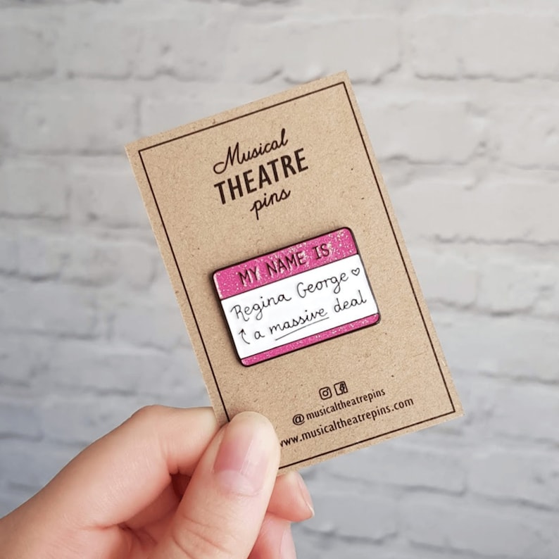 K&ouml;nnte beinhalten: Rosa emaillierte Anstecknadel mit Glitzer, die ein Namensschild mit der Aufschrift "My name is Regina George, a massive deal." zeigt.