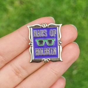 May include: A gold-colored metal pin with a rectangular purple background. The pin features the text "HAUS OF HOLBEIN" in white lettering, with a green glasses graphic. The pin is held in a hand against a green background.
