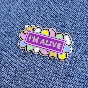 May include: A purple enamel pin with a white border and the text "I'M ALIVE" in white letters. The pin is surrounded by colourful shapes, including stars, pills, and a crescent moon.