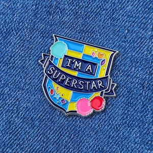 以下が含まれることがあります： 白いバナーに「I'm a Superstar」と書かれた、盾の形をしたカラフルなエナメルピン。盾は青と黄色の背景にカラフルなアクセントが施されています。