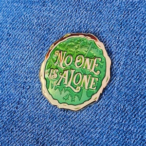 May include: A circular enamel pin with a gold edge, featuring the text "No One Is Alone" in green and gold. The pin has a green background with a forest design.