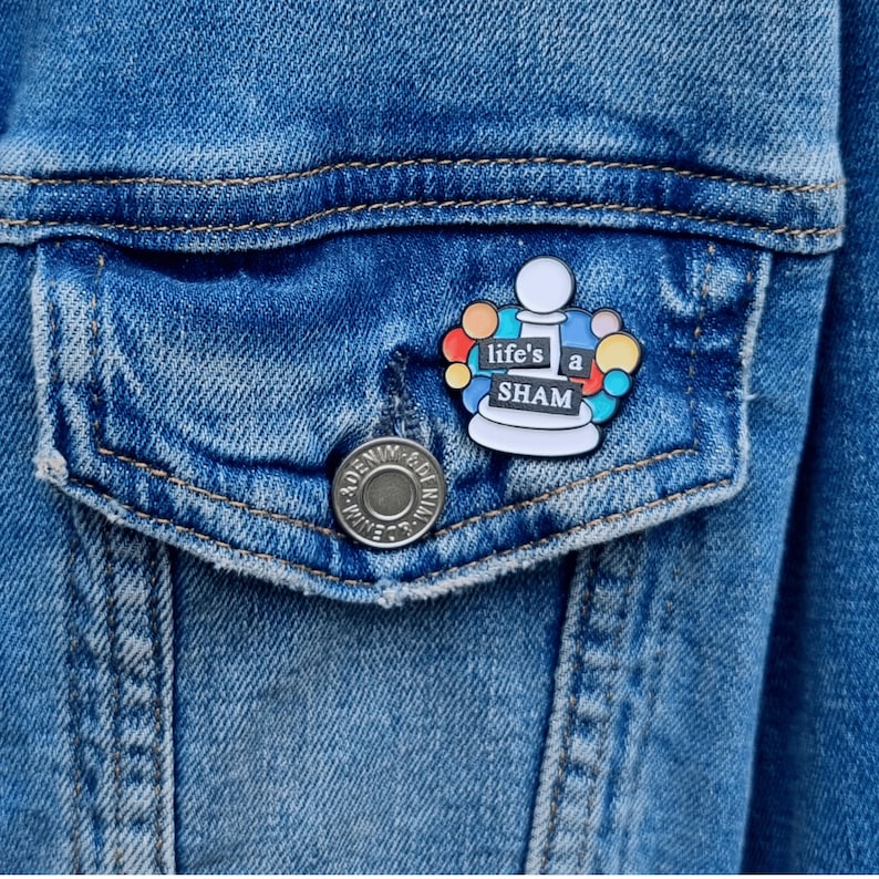May include: A colorful enamel pin featuring a white chess piece with the text "life's a sham" in black lettering. The chess piece is surrounded by colorful circles.