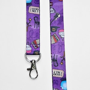 Peut inclure: Lanière violette avec un motif répété d'instruments de musique, de cupcakes, de chaises et d'étoiles. La lanière est munie d'un mousqueton pivotant argenté. Le texte "Lucky No. 7" est répété sur la lanière.