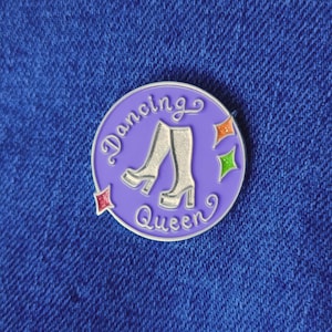 Könnte beinhalten: Eine lilafarbene Emaille-Anstecknadel mit silbernem Rand. Die Anstecknadel zeigt ein Paar weiße Stiefel mit silbernem Rand und den Text "Dancing Queen" in silberner Kursivschrift. Außerdem befinden sich drei kleine, bunte Diamanten um die Stiefel herum.