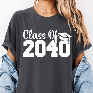 May include: A charcoal gray short-sleeved t-shirt featuring the white printed text Class Of 2040 with a graphic of a graduation cap. The shirt has a crew neckline and a relaxed fit, layered under a light blue denim jacket with visible buttons.
