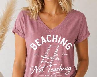May include: A mauve V-neck t-shirt with rolled sleeves. The shirt features the white text "BEACHING Not Teaching" with a beach chair graphic. The text also includes "EST. 2026" and "#RETIRED". The model is wearing blue jeans.