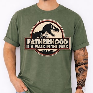 May include: Moss green t-shirt with a graphic featuring a dinosaur silhouette and the text "FATHERHOOD IS A WALK IN THE PARK" in a circular design. The shirt has short sleeves and a relaxed fit.