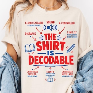 May include: An ivory t-shirt featuring red and blue text that reads THE SHIRT IS DECODABLE, surrounded by educational linguistic labels including CLOSED SYLLABLE, R-CONTROLLED, DIGRAPHS, and icons of a speaker, an open book, and a pencil.