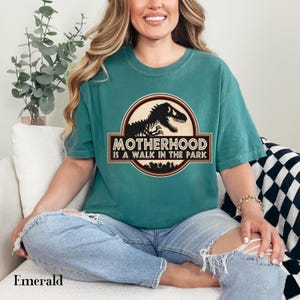 May include: Emerald green t-shirt with a graphic featuring a dinosaur silhouette and the text "MOTHERHOOD IS A WALK IN THE PARK." The shirt is a crew neck and has a relaxed fit. The word "Emerald" is printed at the bottom.