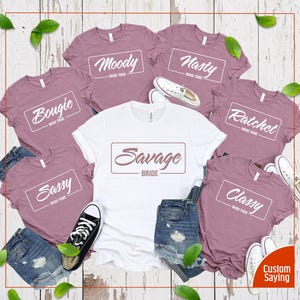 May include: Six t-shirts with different text on each. The shirts are mauve and white. The text on the shirts says "Bougie Bride Tribe", "Moody Bride Tribe", "Nasty Bride Tribe", "Ratchet Bride Tribe", "Sassy Bride Tribe", and "Classy Bride Tribe". One shirt says "Savage Bride" in a white rectangle with a brown outline.