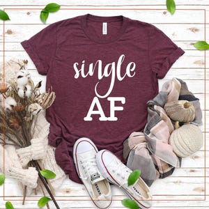 May include: Maroon t-shirt with the words "single AF" in white script. The shirt is paired with white canvas sneakers, a patterned scarf, and decorative elements on a wooden background. The shirt is a casual wear item.