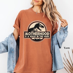 May include: A burnt orange t-shirt with a graphic that reads "MOTHERHOOD IS A WALK IN THE PARK." The graphic features a black dinosaur silhouette inside a circular emblem with a black and white border. The shirt has rolled-up sleeves.