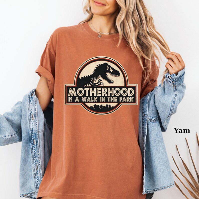 May include: A burnt orange t-shirt with a graphic that reads "MOTHERHOOD IS A WALK IN THE PARK." The graphic features a black dinosaur silhouette inside a circular emblem with a black and white border. The shirt has rolled-up sleeves.