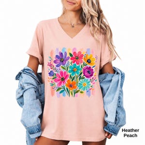 Op de afbeelding: Een perzikkleurig T-shirt met V-hals en een kleurrijke grafische print van een boeket bloemen in paars, roze, geel en turquoise. Het ontwerp is geplaatst tegen een achtergrond van verticale pastelkleurige penseelstreken. De tekst Heather Peach is zichtbaar.