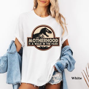 May include: White t-shirt featuring a circular graphic with a dinosaur silhouette and the text "MOTHERHOOD IS A WALK IN THE PARK". The design is in black, brown, and red tones. The shirt is made of a soft, comfortable material.