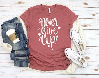 May include: A rust-colored t-shirt with the text "Never give up!" in white lettering with small white stars. The shirt is laid flat on a white wooden surface with a pair of white canvas shoes.