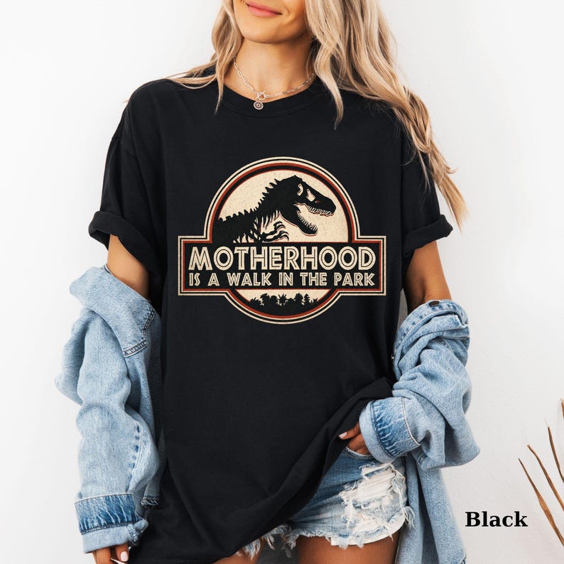 May include: Black t-shirt with a circular graphic featuring a dinosaur silhouette and the text "MOTHERHOOD IS A WALK IN THE PARK". The design is in shades of brown, beige, and black. The shirt is a casual, unisex style.