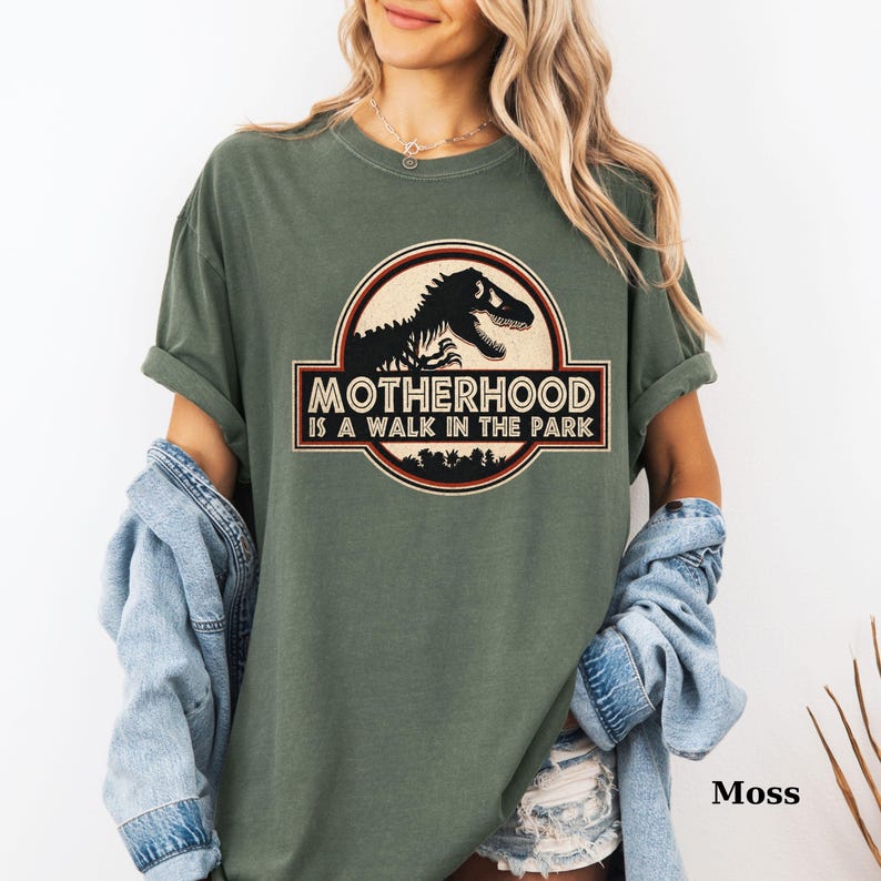 May include: Moss green t-shirt featuring a circular graphic with a dinosaur silhouette and the text "MOTHERHOOD IS A WALK IN THE PARK". The design is in black, red, and cream colors. The shirt has rolled-up sleeves and a relaxed fit.