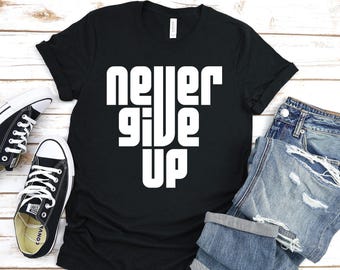May include: Black t-shirt with white text that reads "never give up" in a bold, blocky font. The shirt is laid flat on a white wooden surface with a pair of black and white sneakers and a pair of blue jeans.