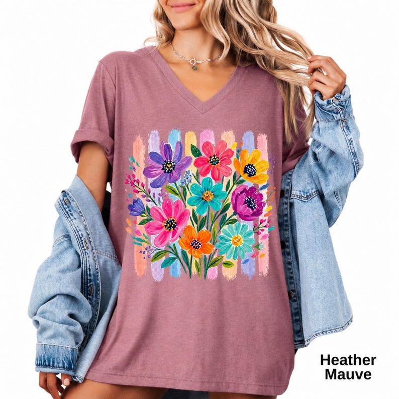 Op de afbeelding: Een heather mauve V-hals t-shirt met een kleurrijke afbeelding van bloemen in roze, paars, geel en groenblauw, geplaatst tegen een achtergrond van verticale pastelkleurige penseelstreken. Een lichtblauw spijkerjack hangt over de schouders.