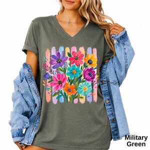 Op de afbeelding: Een militair groen T-shirt met V-hals en een centrale afbeelding van een kleurrijk boeket bloemen in roze, paarse, blauwe en oranje tinten, geplaatst tegen een achtergrond van verticale pastelkleurige penseelstreken. Een blauw spijkerjack wordt over het shirt gedragen.