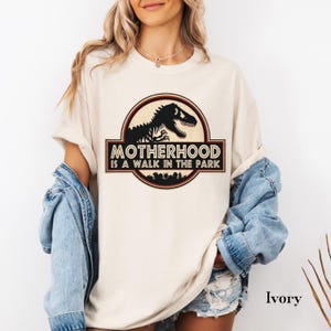 May include: Ivory-colored t-shirt featuring a circular graphic with a black dinosaur silhouette and the text "MOTHERHOOD IS A WALK IN THE PARK." The design is framed by a red and brown border. The shirt has short sleeves.