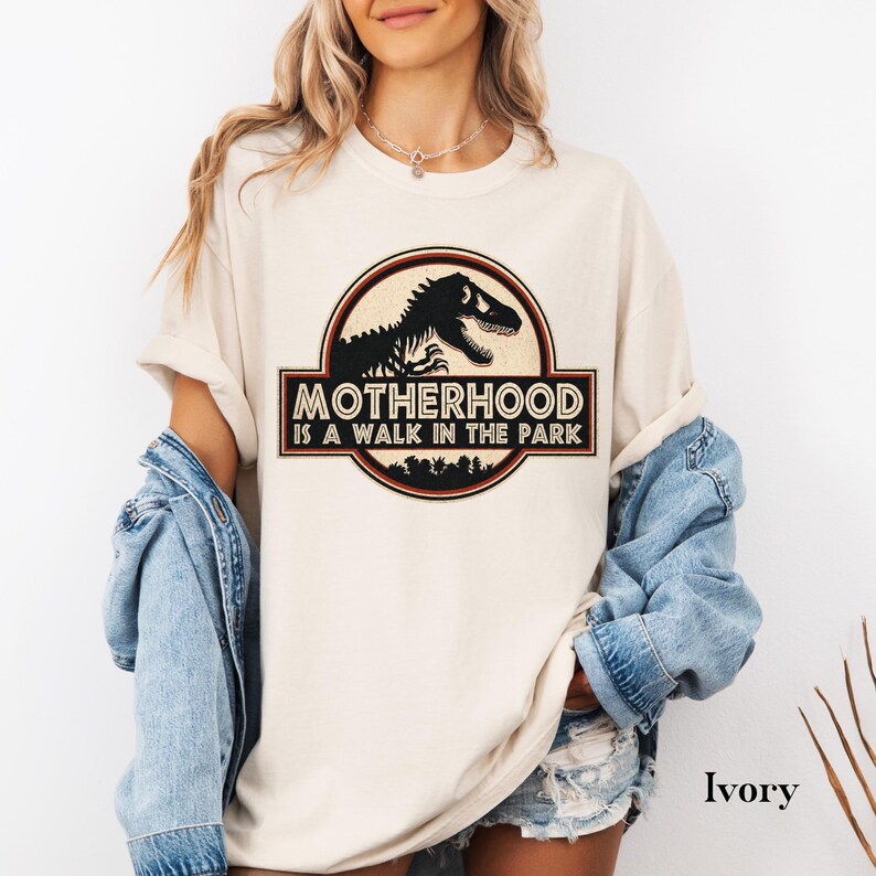 May include: Ivory-colored t-shirt featuring a circular graphic with a black dinosaur silhouette and the text "MOTHERHOOD IS A WALK IN THE PARK." The design is framed by a red and brown border. The shirt has short sleeves.