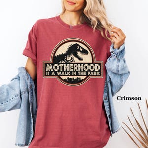 May include: Crimson-colored t-shirt featuring a circular graphic with a dinosaur silhouette and the text "MOTHERHOOD IS A WALK IN THE PARK." The design includes a forest silhouette at the bottom. The shirt has short sleeves and a relaxed fit.