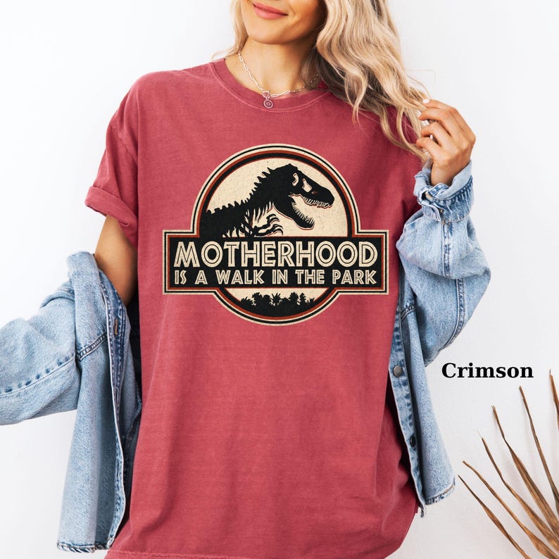 May include: Crimson-colored t-shirt featuring a circular graphic with a dinosaur silhouette and the text "MOTHERHOOD IS A WALK IN THE PARK." The design includes a forest silhouette at the bottom. The shirt has short sleeves and a relaxed fit.