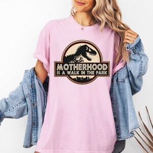 May include: A light pink t-shirt with the text "MOTHERHOOD IS A WALK IN THE PARK" in a black and brown graphic. The graphic features a dinosaur silhouette inside a circular frame.
